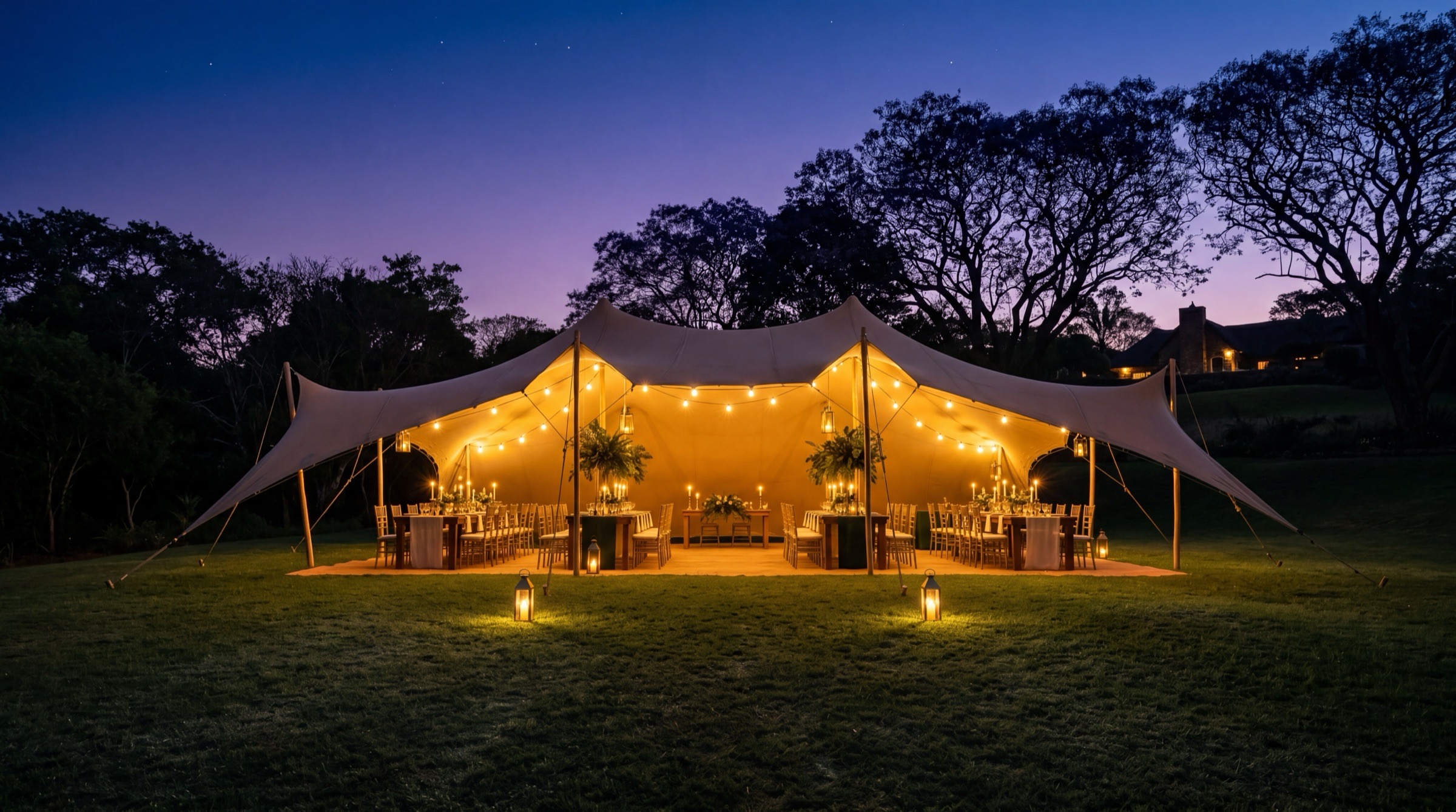 A Heritage stretch tent glowing warmly from within on a manicured lawn at twilight, brass bistro lights illuminating dressed tables, jacaranda trees silhouetted against the dusk-blue African sky